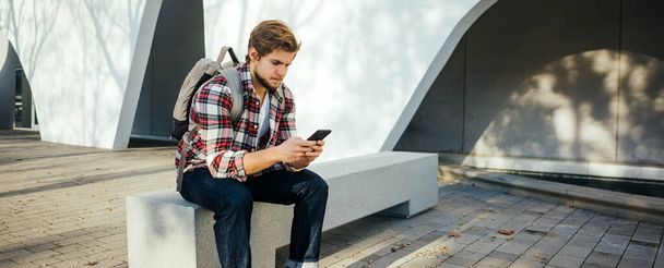 Retrato de un joven moderno con smartphone en una calle. Hipster chico con móvil en su mano al aire libre - Foto, Imagen