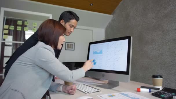 Μεγέθυνση της Ασιάτισσας επιχειρηματία δείχνοντας με στυλό στην οθόνη του υπολογιστή και εξηγώντας διάγραμμα στον άνδρα συνάδελφό της που στέκεται κοντά - Πλάνα, βίντεο