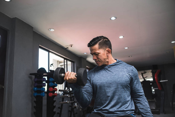A fit asian man in a sweatshirt does alternating seated dumbbell curls. Working out and training biceps. Open air gym setting. - Photo, Image