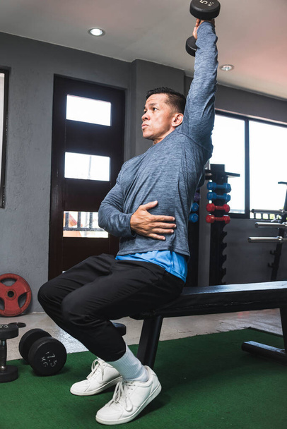 A fit athletic asian guy in a sweatshirt does seated overhead dumbbell one arm tricep extensions. Working out and training triceps. Open air gym setting. - Photo, Image
