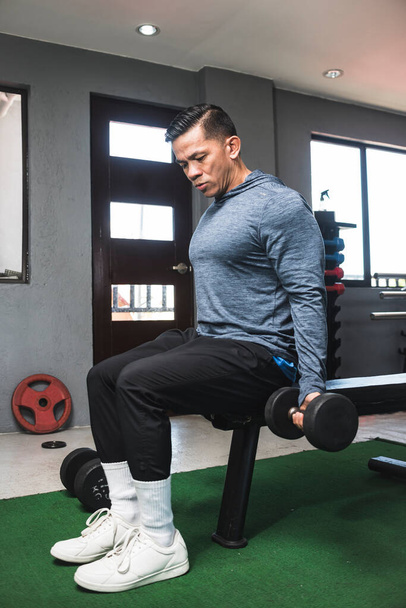 A fit athletic man in a sweatshirt does seated dumbbell curls. Working out and training biceps. Open air gym setting. - Foto, Bild