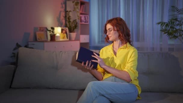 femme avec des lunettes lit un livre intéressant et lève les yeux de rêve tout en se relaxant à la maison sur le canapé la nuit - Séquence, vidéo