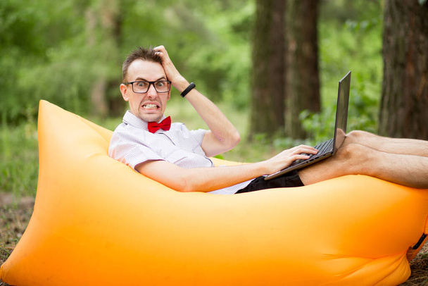 Verängstigter Student mit Brille und Laptop auf aufblasbarer Matratze im Sommerpark. Bildung, Wirtschaft, Scheitern und Technologiekonzept - Foto, Bild