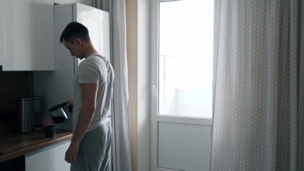 Schöner junger Mann gießt Kaffee aus Kaffeemaschine und geht zum Fenster in der Küche - Filmmaterial, Video