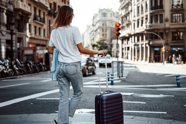 Νεαρή γυναίκα που ταξιδεύει με αποσκευές σε ένα ηλιόλουστο δρόμο της πόλης. Ταξιδιώτης σε διακοπές. Αναμονή για ταξί - Φωτογραφία, εικόνα