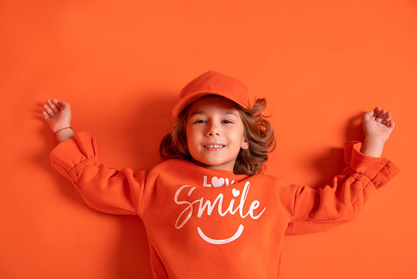 Χαμογελαστό κοριτσάκι 6-7 ετών με καπέλο και μοντέρνα ρούχα βρίσκεται στο πάτωμα του στούντιο σε πορτοκαλί φόντο. Αντιγραφή χώρου - Φωτογραφία, εικόνα