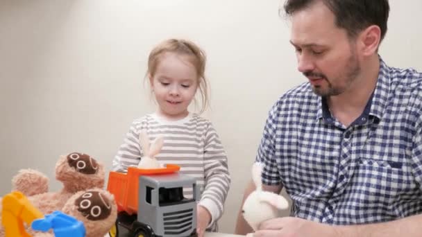 Caring father cheerfully helping cute child to drive toy truck, enjoy tgame. Happy family daughter and dad play toy cars and plush animals of a hare and a bear. Parent dad and child play toys at home. - Footage, Video
