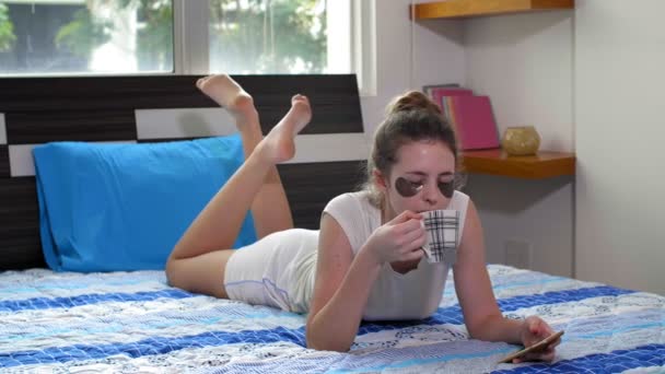 Amplia toma de mujer caucásica joven acostada en la cama con parches debajo de los ojos en su cara, bebiendo té caliente y usando su teléfono - Imágenes, Vídeo
