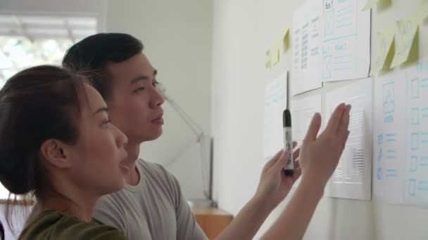Close up of Asian young woman standing at whiteboard and explaining charts to her male business partner standing nearby and listening to her - Footage, Video