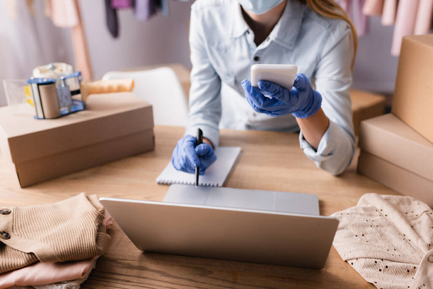częściowy widok właściciela w rękawiczkach lateksowych pracujących ze smartfonem i laptopem w salonie, rozmyte tło - Zdjęcie, obraz