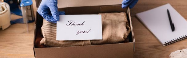 обрезанный вид продавца в латексных перчатках держа карточку с благодарностью письма возле коробки с одеждой, баннер - Фото, изображение