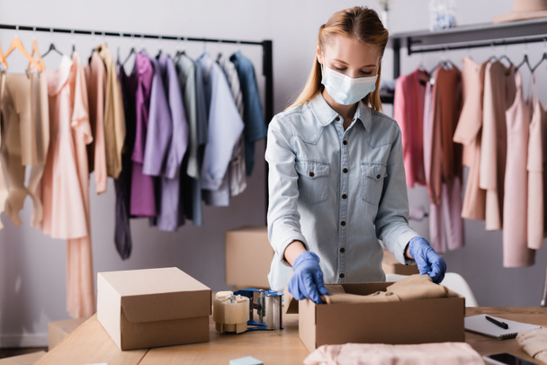 jonge zakenvrouw met medisch masker, kleding inpakken in dozen in de showroom, wazig voorgrond - Foto, afbeelding