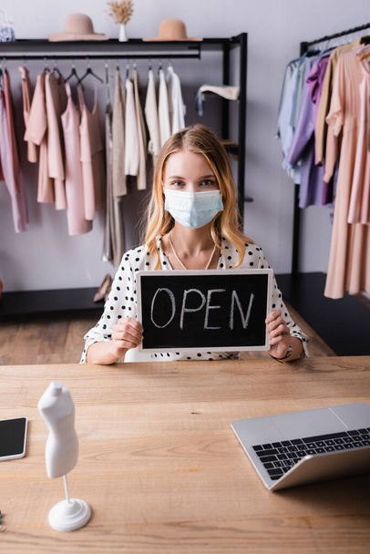 showroom owner in medical mask, holding board with open lettering while sitting at workplace - Photo, Image