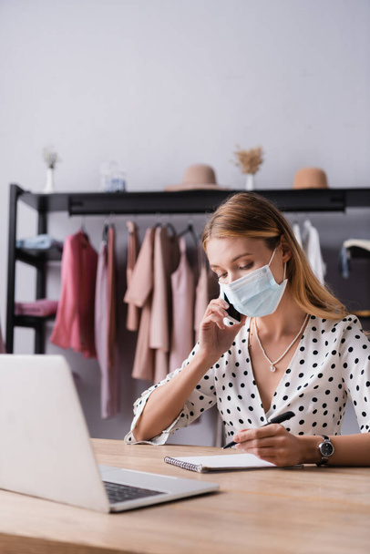 businesswoman in medical mask, talking on smartphone near laptop in showroom - Photo, Image