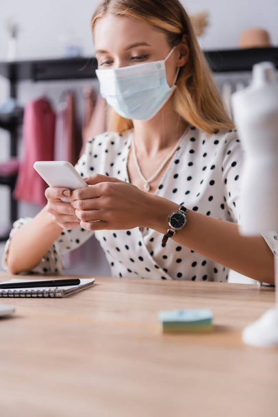 showroom owner in protective mask, messaging on smartphone at workplace on blurred foreground - Photo, Image