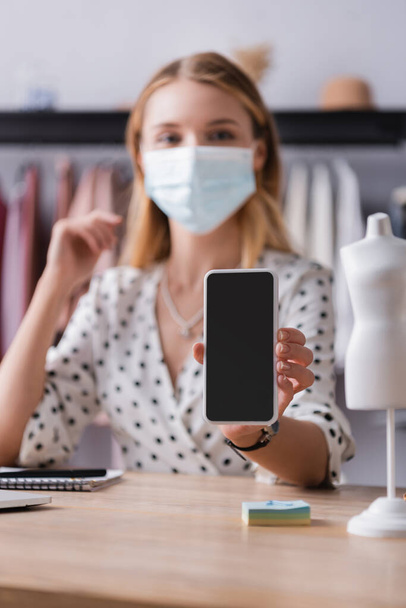 propietario de la sala de exposición en máscara médica, mostrando teléfono inteligente con pantalla en blanco sobre fondo borroso - Foto, imagen