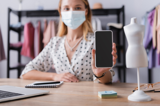 mujer de negocios en máscara médica, mostrando el teléfono celular con pantalla en blanco mientras trabaja en la sala de exposición sobre fondo borroso - Foto, Imagen