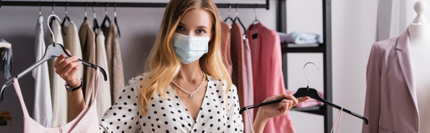 young businesswoman in medical mask, holding hangers and looking at camera, banner - Zdjęcie, obraz