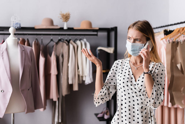 upset showroom proprietor in medical mask talking on mobile phone near mannequin and hangers on blurred background - Photo, Image