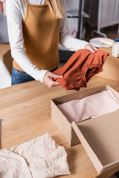 μερική άποψη του ιδιοκτήτη κρατώντας πουλόβερ κοντά κουτί με ρούχα σε εκθεσιακό χώρο - Φωτογραφία, εικόνα