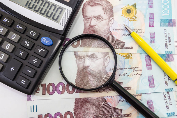 1000 hryvnia con una calculadora y una lupa sobre fondo blanco. Dinero ucraniano. Concepto financiero. - Foto, imagen
