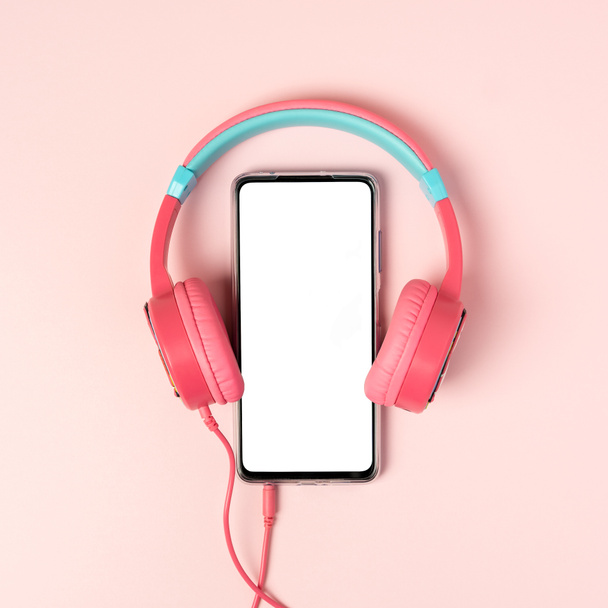 Una vista dall'alto sullo smartphone con schermo bianco e auricolari su sfondo rosa. Studio girato concetto di comunicazione e musica. Modello finto, disposizione piatta, spazio di copia. - Foto, immagini