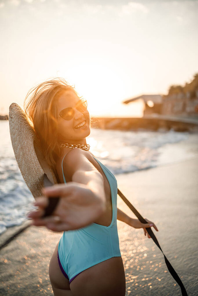 Μια γυναίκα με μαγιό και ένα καπέλο με γυαλιά περπατά κατά μήκος της παραλίας το ηλιοβασίλεμα. Η έννοια της θαλάσσιας αναψυχής. Επιλεκτική εστίαση - Φωτογραφία, εικόνα