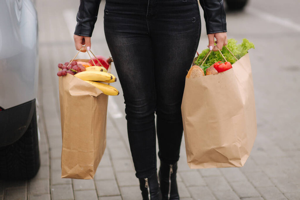 Zakupy z supermarketu w opakowaniu z eko-rzemiosłem. Dostawa żywności podczas kwarantanny. Papierowe eko-torby pełne świeżego jedzenia. Kobieta trzyma dwie paczki przy samochodzie. Zbliżenie - Zdjęcie, obraz