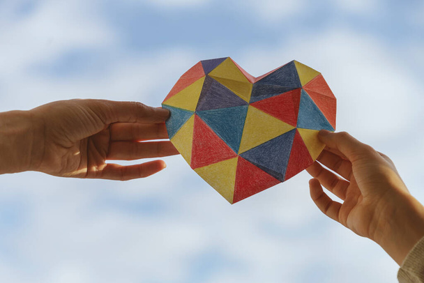 Взрослые и дети держат в руках красочное бумажное сердце на голубом фоне неба. Аутистический спектр расстройства семейной поддержки. Концепция Всемирного дня борьбы с аутизмом - Фото, изображение