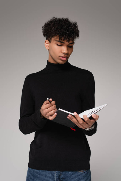 joven afroamericano en jersey de cuello alto sosteniendo cuaderno y bolígrafo aislado en gris - Foto, imagen