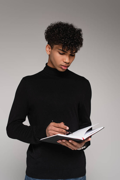 joven afroamericano en jersey de cuello alto escrito en cuaderno aislado en gris - Foto, imagen