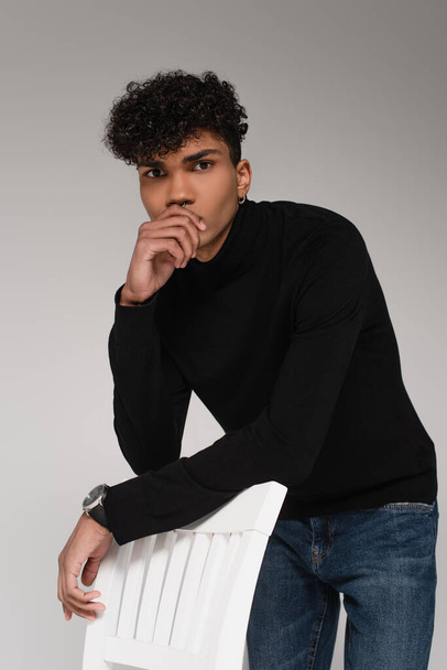 young african american man in turtleneck sweater posing near chair isolated on grey - Foto, imagen