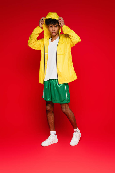 longitud completa de hombre afroamericano con estilo en chaqueta de lluvia amarilla ajustando la capucha en rojo - Foto, Imagen