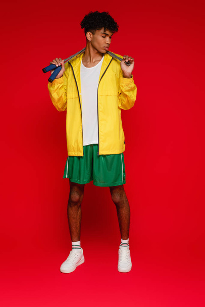 full length of sportive african american man in yellow rain jacket standing with skipping rope on red - Fotoğraf, Görsel