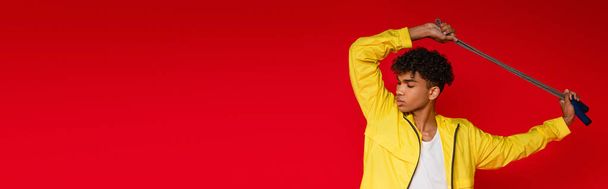 uomo afroamericano trafitto in giacca gialla in posa con corda da salto isolata su rosso, stendardo - Foto, immagini
