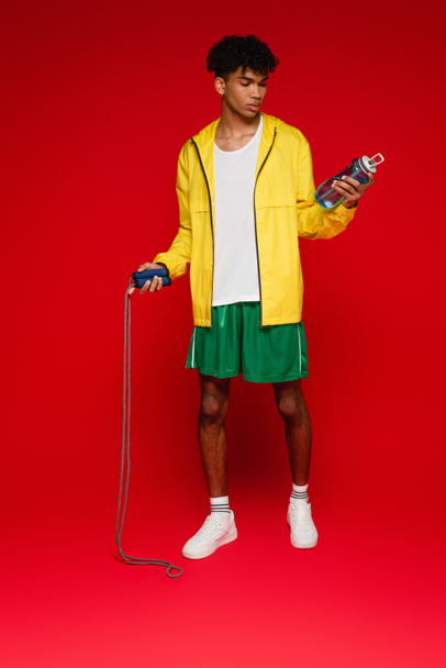 homme afro-américain sportif en veste de pluie jaune debout avec corde à sauter et bouteille de sport sur rouge - Photo, image
