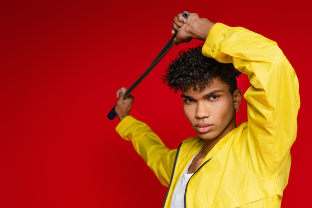 pierced african american man in yellow jacket posing with jumping rope isolated on red - Photo, Image