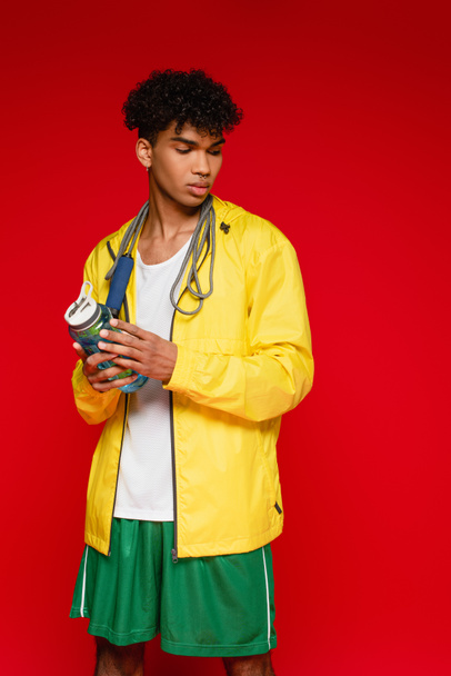 curly african american man in yellow rain jacket standing with skipping rope and sports bottle isolated on red - Foto, Imagem