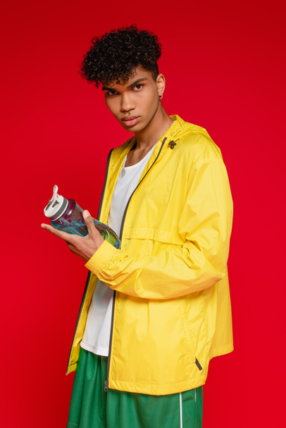 curly african american man in yellow rain jacket standing with sports bottle isolated on red - Photo, Image