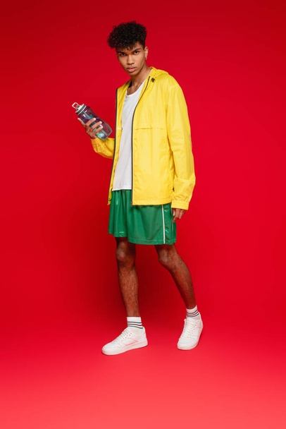 full length of curly african american man in yellow rain jacket standing with sports bottle on red - Foto, Imagem