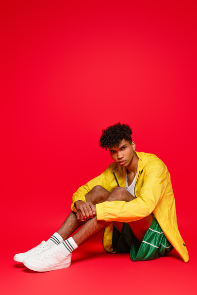full length of curly african american man in yellow jacket sitting while posing on red - Foto, imagen