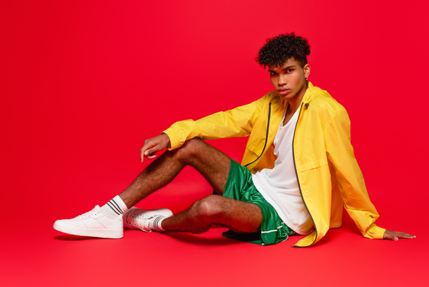 full length of pierced african american man in yellow jacket and white sneakers sitting while posing on red - Фото, изображение