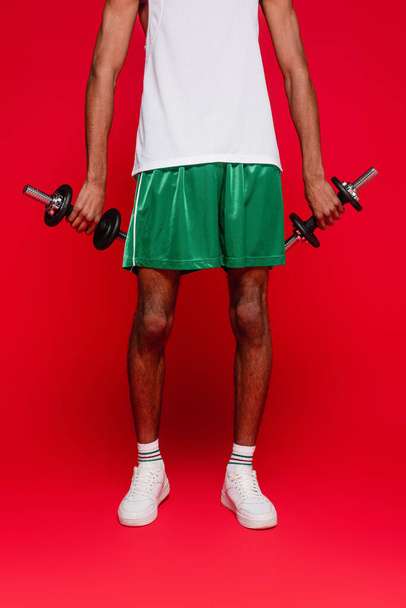 recortado vista de deportivo africano americano hombre en pantalones cortos y tanque de ejercicio superior con mancuernas en rojo - Foto, imagen