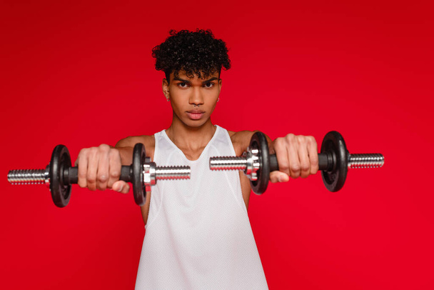 sportive african american man in tank top exercising with dumbbells on blurred foreground isolated on red - Photo, Image