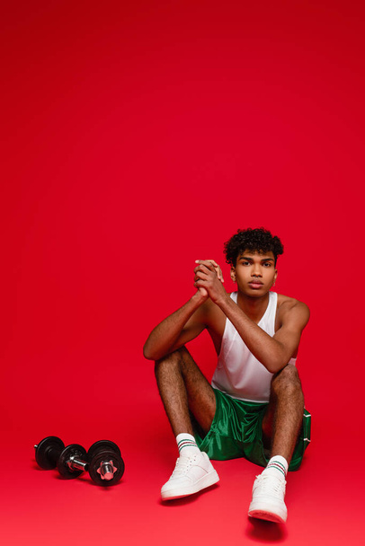 sportive african american man in tank top and shorts sitting near dumbbells on red - Foto, imagen