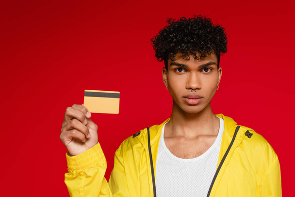 perforado africano americano hombre en chaqueta celebración de tarjeta de crédito aislado en rojo - Foto, imagen