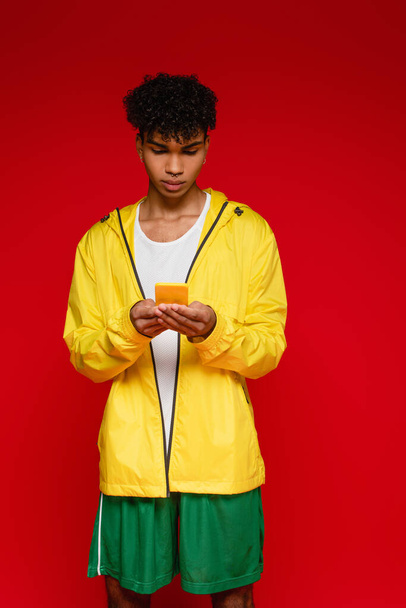 curly african american man in jacket using smartphone isolated on red - Photo, Image