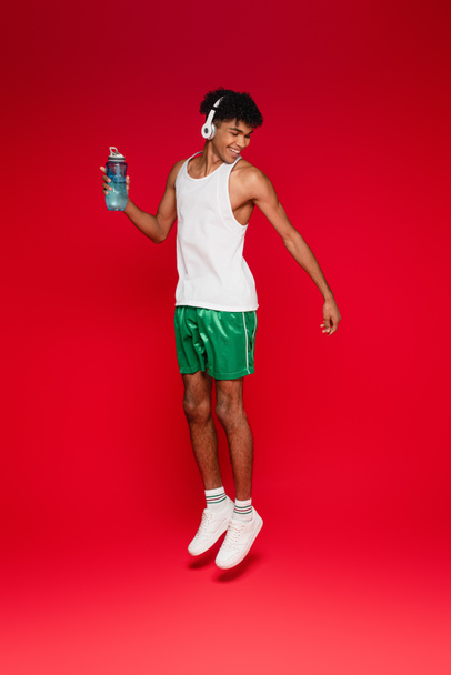pleine longueur de sourire homme afro-américain en casque sans fil tenant bouteille de sport tout en sautant sur rouge - Photo, image