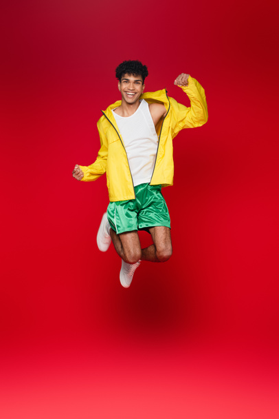повна довжина усміхненого афроамериканця в жовтій куртці, що стрибає на червоному
 - Фото, зображення