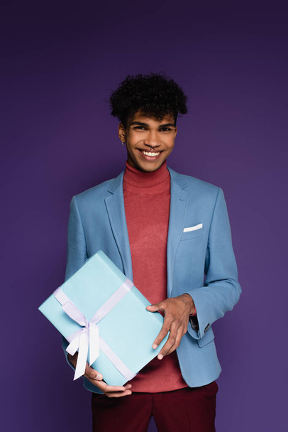 happy african american man in blue blazer holding wrapped present on purple - Фото, изображение